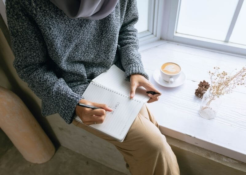 窓際でコーヒーを飲みながら女性ノートに文字を書く女性